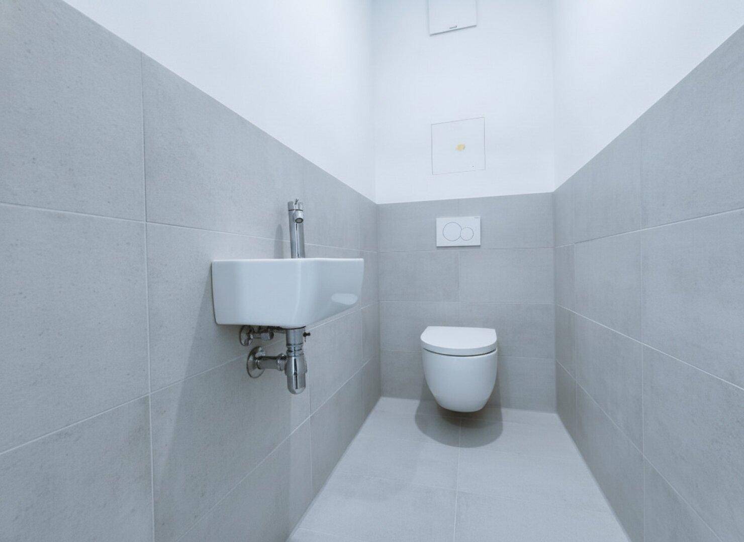 Modernes Gäste-WC mit Wand-WC, kleinem Waschbecken und grauen Wandfliesen.