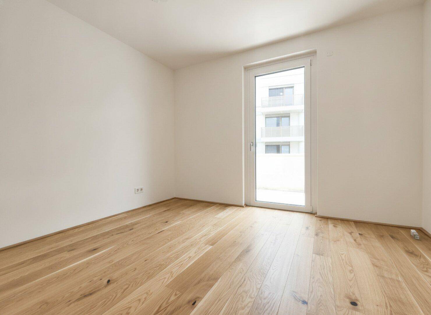 Freundliches Zimmer mit Parkettboden und bodentiefem Fenster, ideal als Schlaf- oder Arbeitszimmer nutzbar.