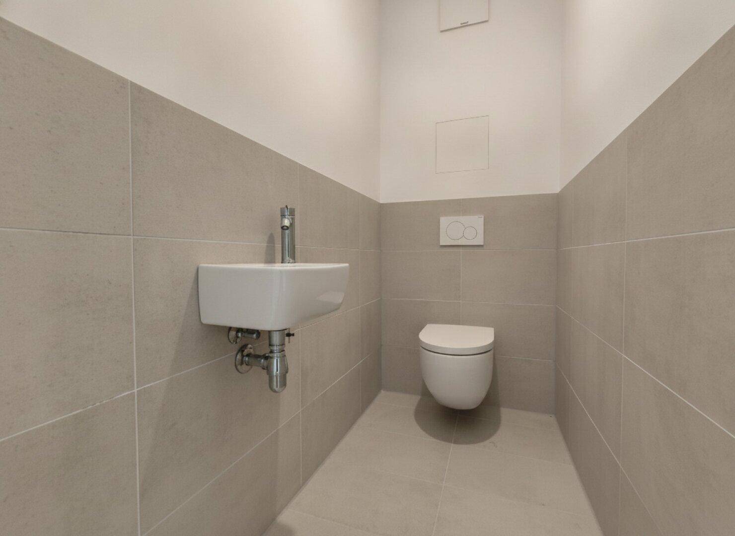 Separates Gäste-WC mit modernem Wand-WC und kleinem Waschbecken, halbhoch gefliest in neutralen Tönen.