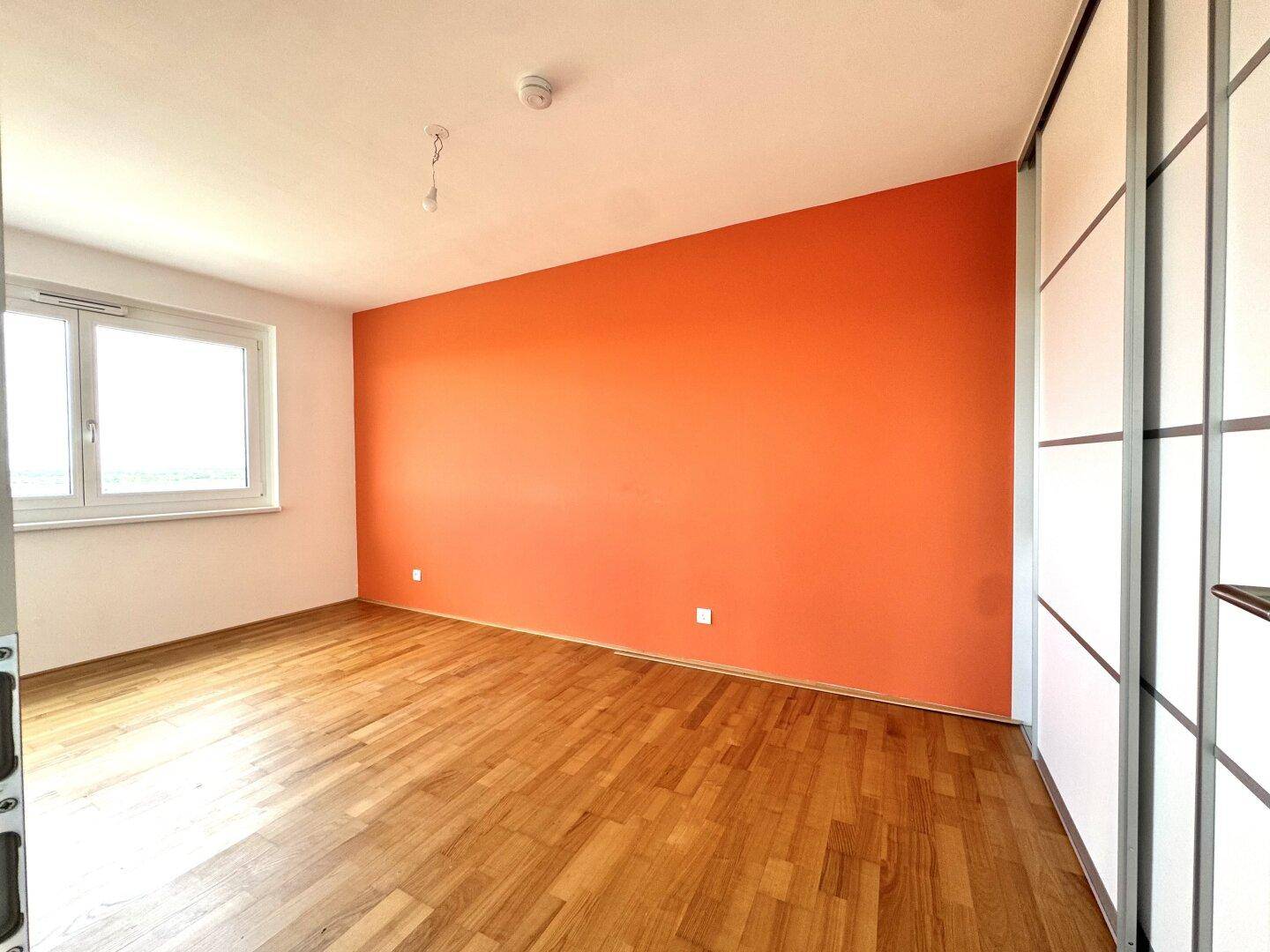Helles Schlafzimmer mit Parkettboden, orangefarbener Wand und Fenster mit Ausblick.