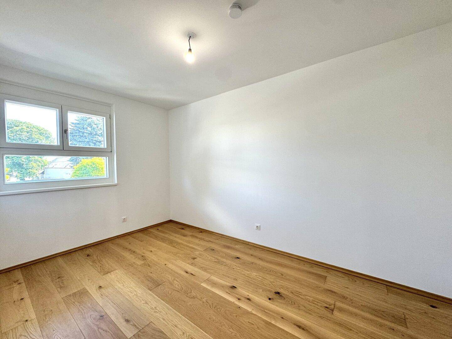 Leeres Zimmer mit hellem Holzfußboden und Fenster mit Blick ins Grüne.