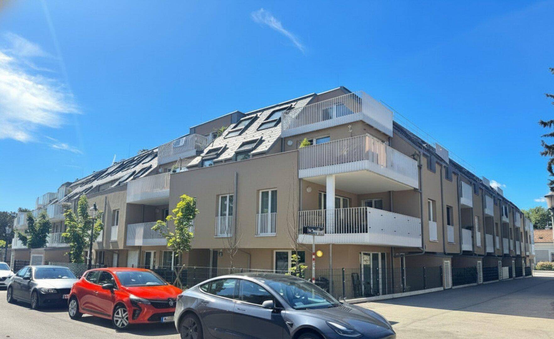 Modernes Mehrfamilienhaus mit beigefarbener Fassade, Balkonen und Mansarddach unter blauem Himmel.