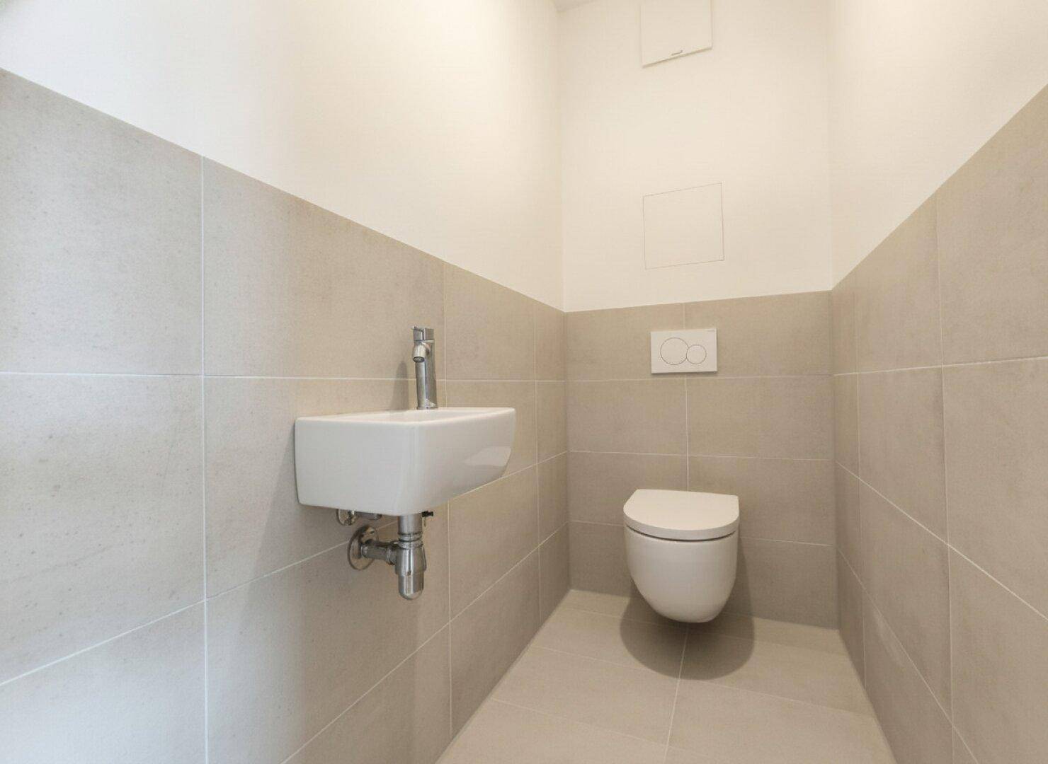 Funktionale Gästetoilette mit wandhängendem WC und kleinem Waschbecken, halbhoch gefliest.