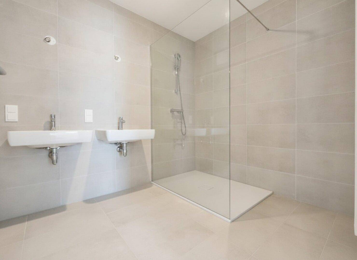 Modernes Badezimmer mit zwei Waschbecken und einer ebenerdigen Dusche, gefliest in hellen Grautönen.
