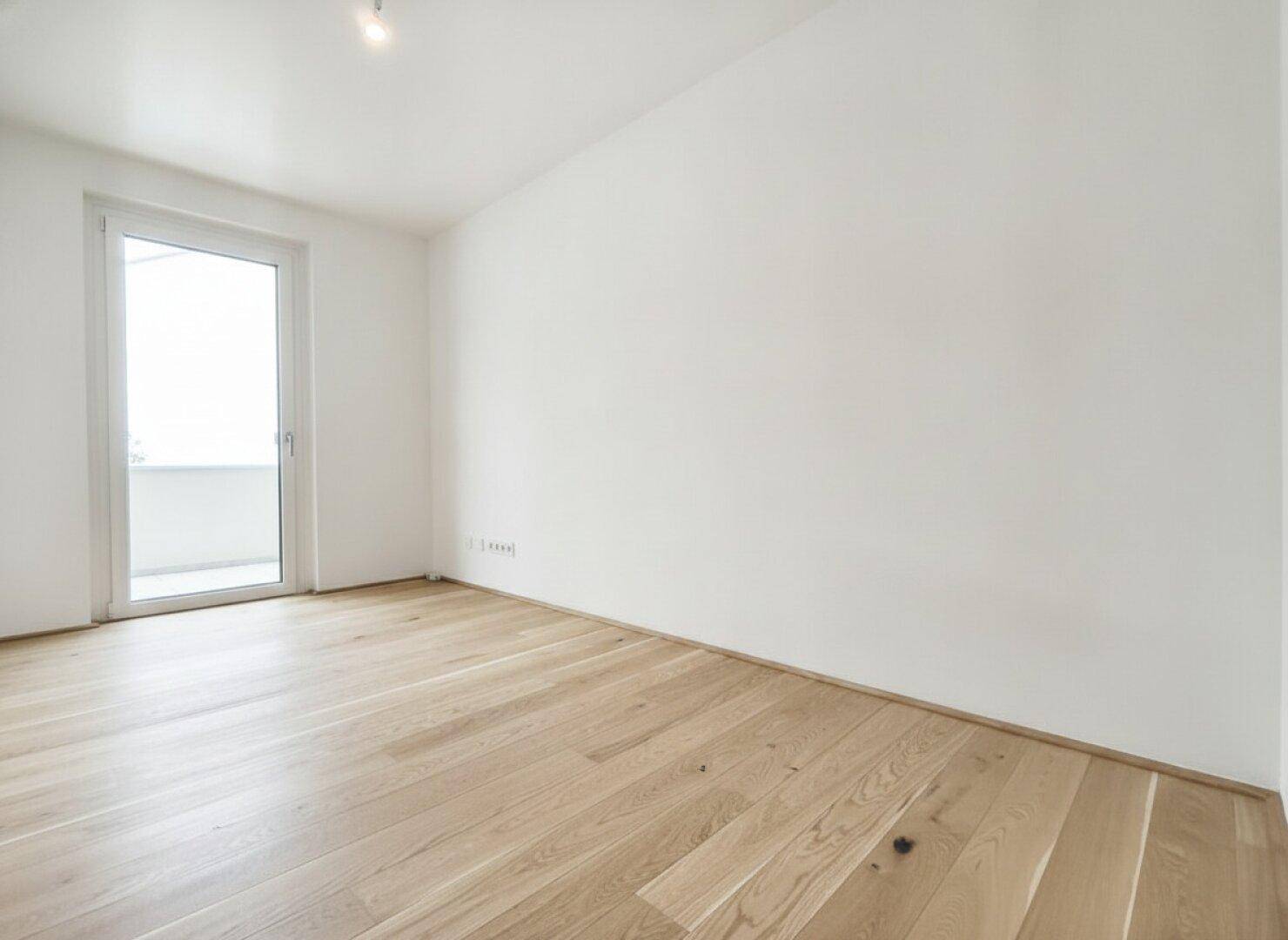 Leeres Schlafzimmer mit hellem Parkettboden und Zugang zu einem kleinen Balkon, ideal für natürliches Licht.