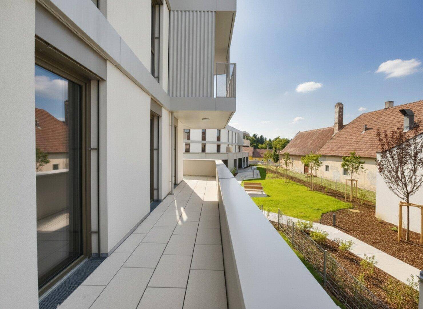 Langer Balkon mit Fliesenboden und seitlichem Blick auf die Wohnanlage und grüne Umgebung.