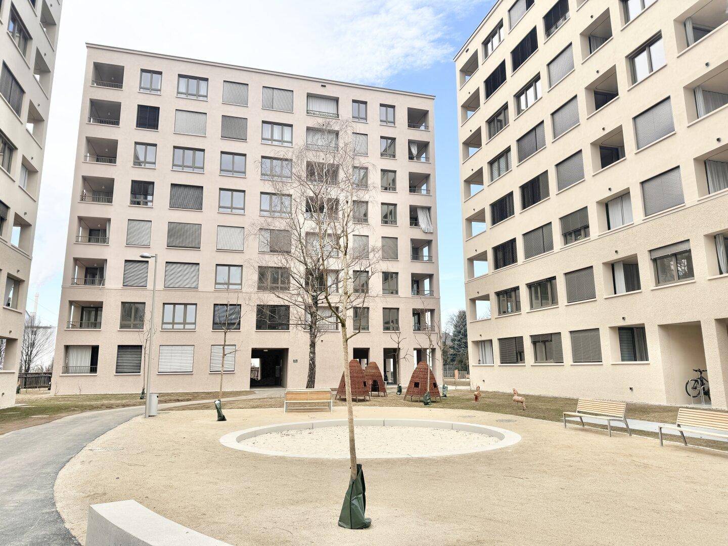 Außenbereich eines Wohnkomplexes mit Spielplatz, Sitzbänken und modernen Gebäuden unter blauem Himmel.
