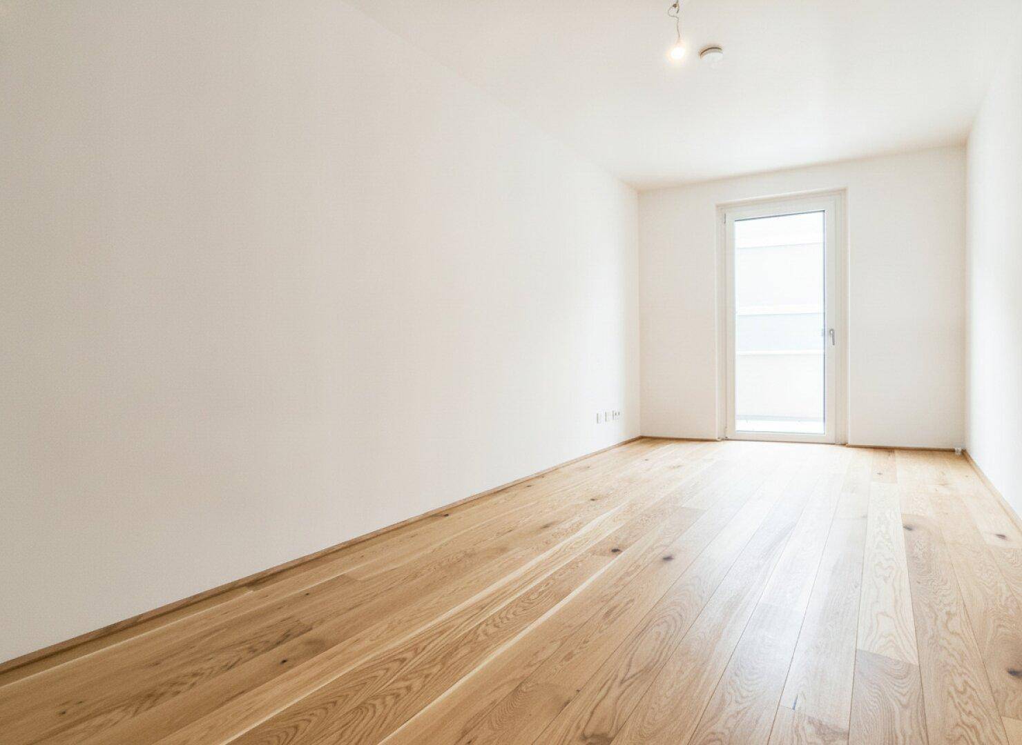 Helles, leeres Zimmer mit Holzboden und Balkontür, bietet viel Potenzial für individuelle Gestaltung.