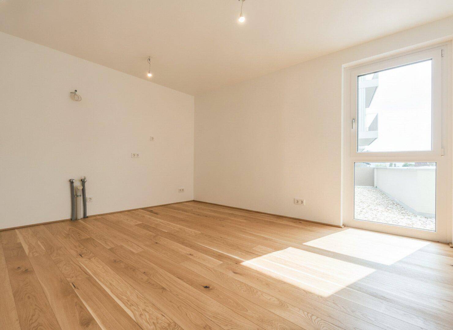 Heller Raum mit Holzboden und einem großen Fenster, das auf eine Terrasse oder einen Balkon führt.