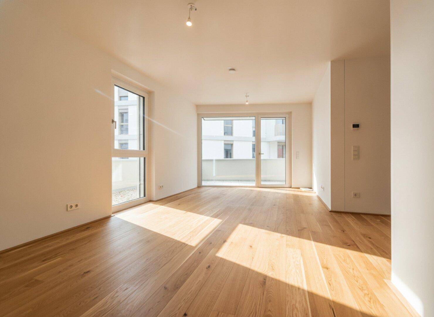 Geräumiger, heller Wohnraum mit Holzboden und großen Fenstern, die viel Tageslicht hereinlassen.