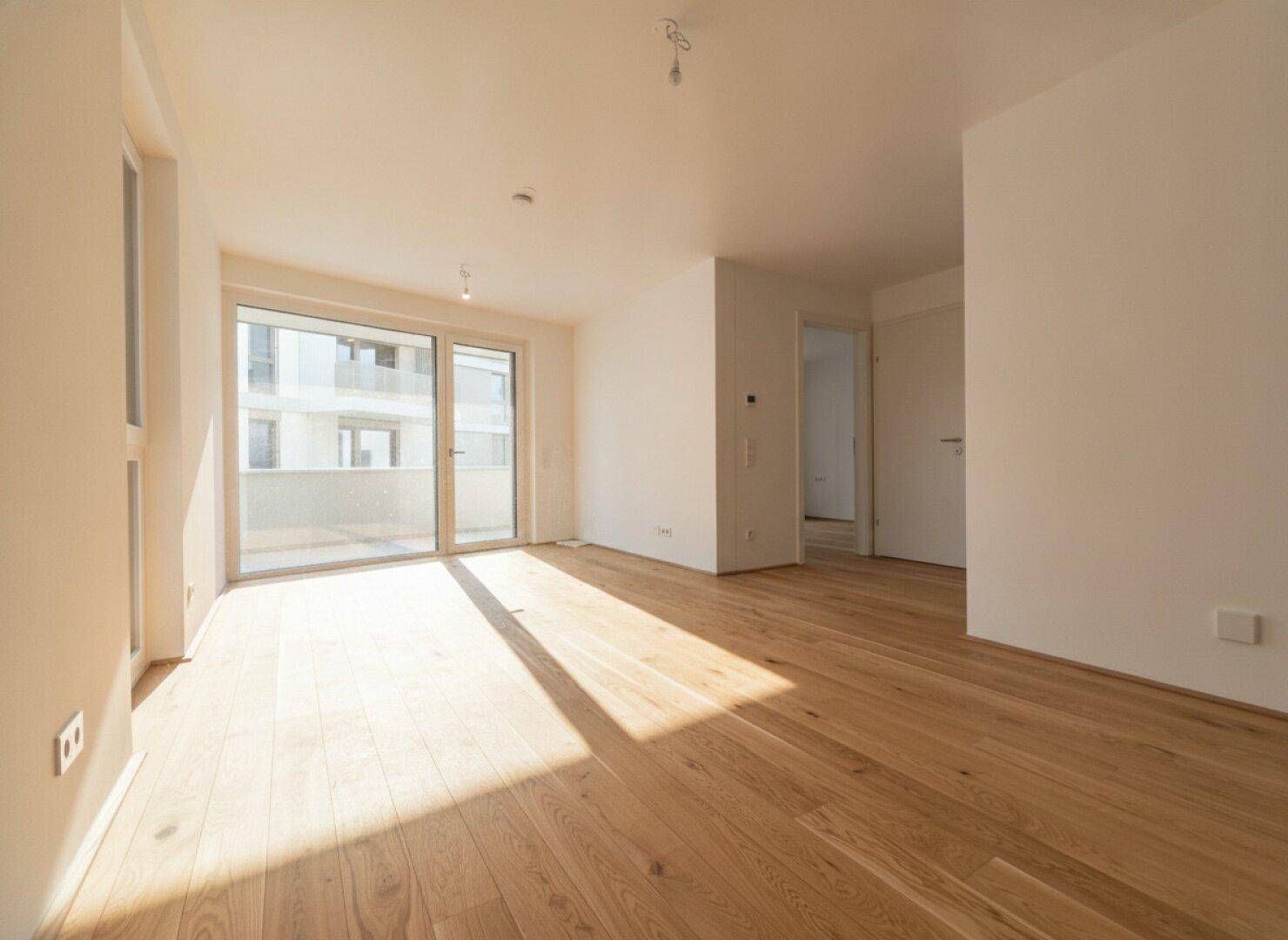 Lichtdurchfluteter Wohnbereich mit hellem Parkettboden und Zugang zu einem Balkon.