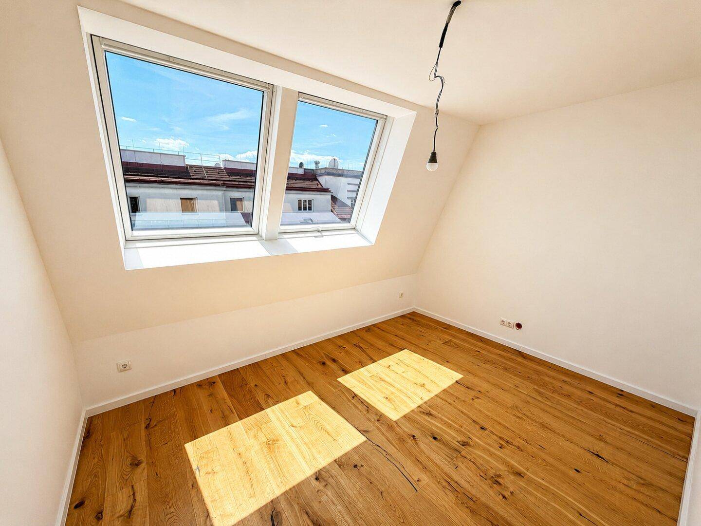 Sonniger Raum mit Holzboden, Dachschrägen und zwei Dachfenstern, die viel Licht hereinlassen.