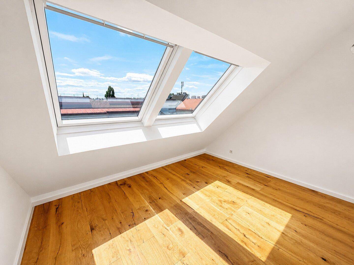 Sonniger Raum mit Holzboden, Dachschrägen und zwei Dachfenstern, die den Raum erhellen.