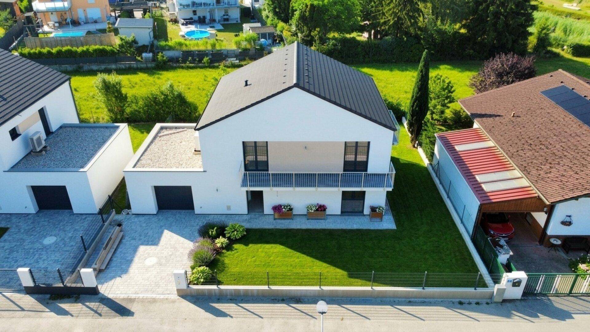 Moderne Neubauvilla mit weißer Fassade, dunklem Dach und gepflegtem Garten in grüner Umgebung.