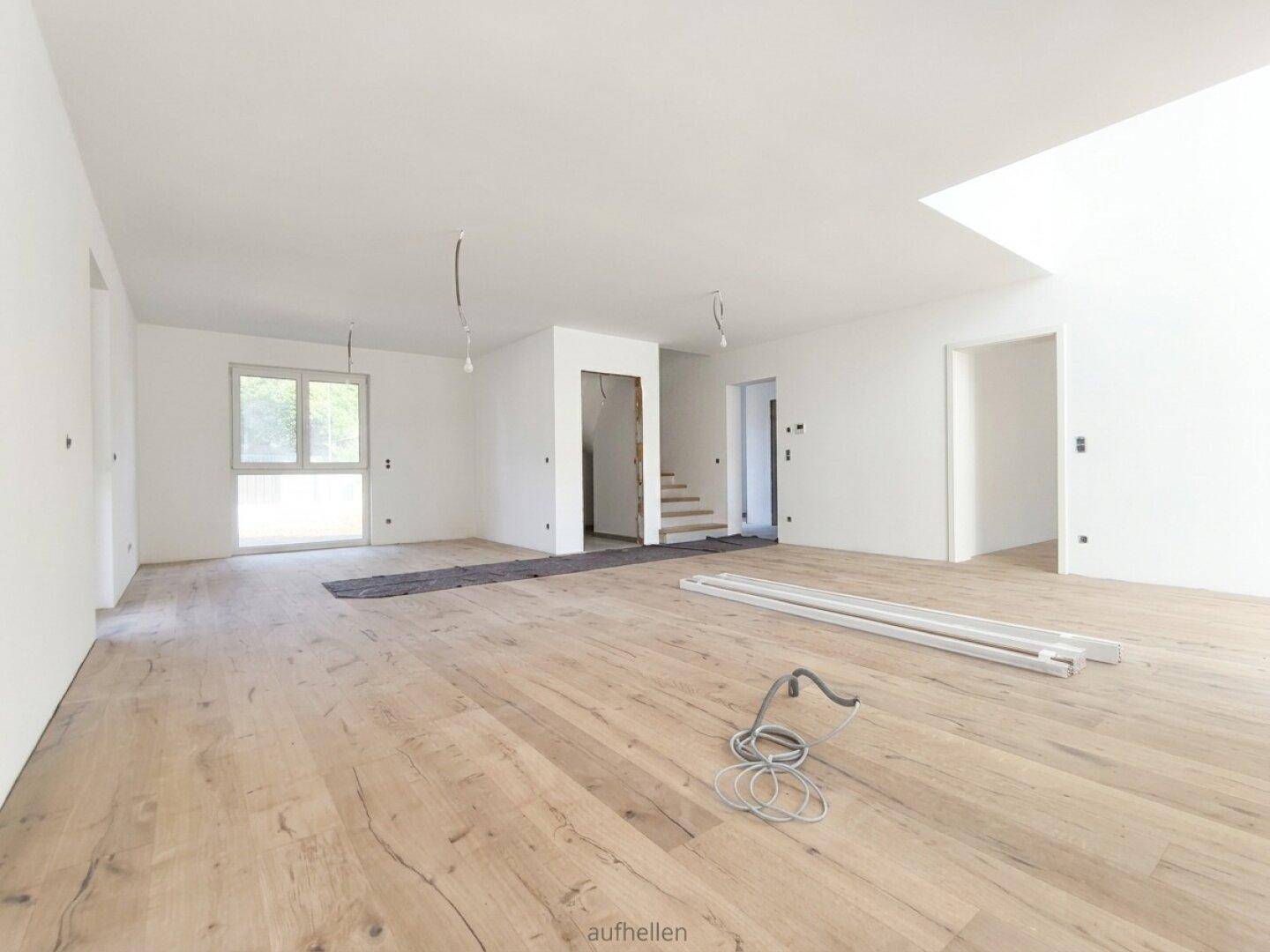 Geräumiger, unmöblierter Wohnbereich mit Holzboden, weißen Wänden und offenem Treppenaufgang.