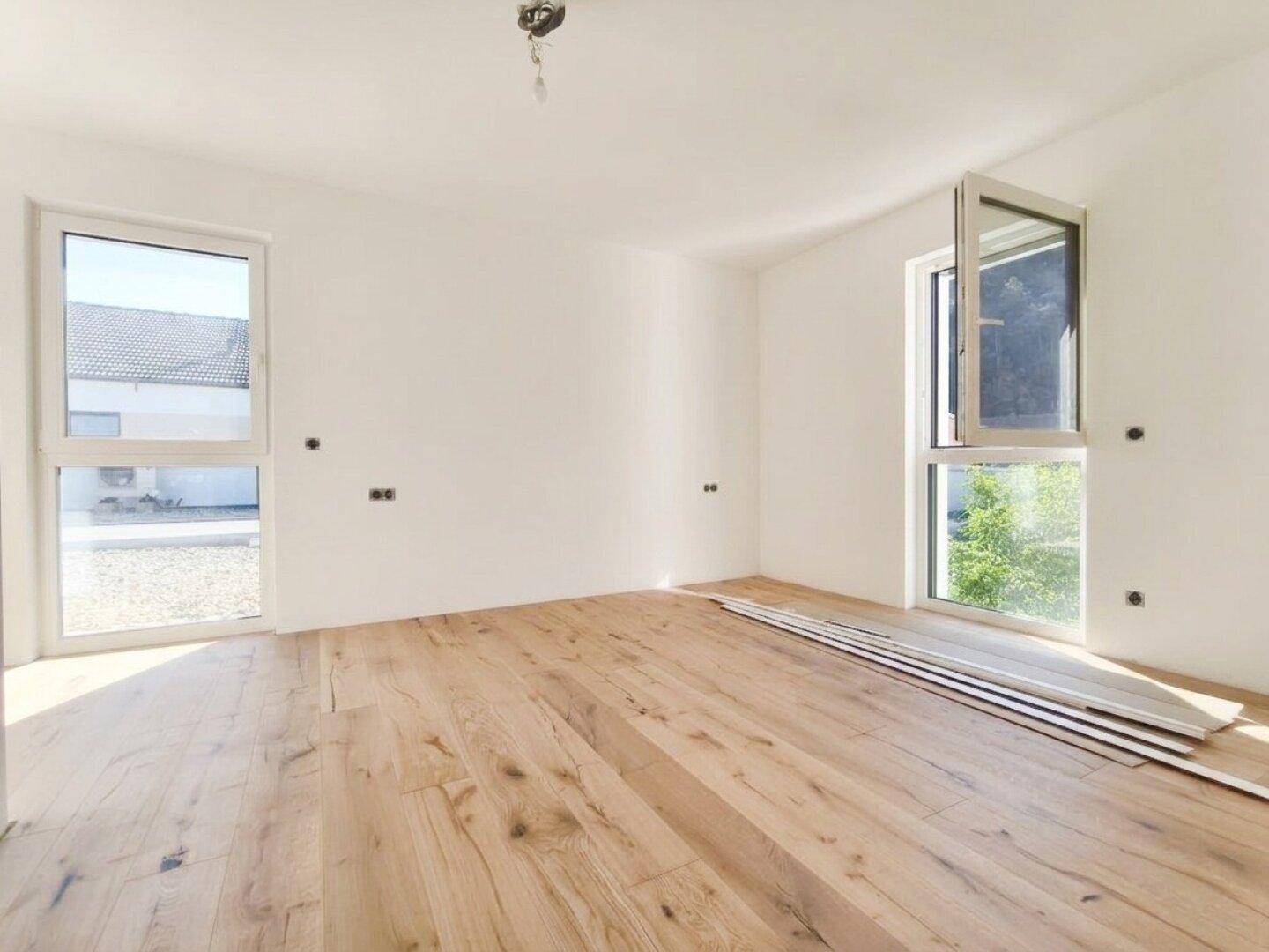 Helles Schlafzimmer mit Holzboden und zwei großen Fenstern, die viel Tageslicht hereinlassen.
