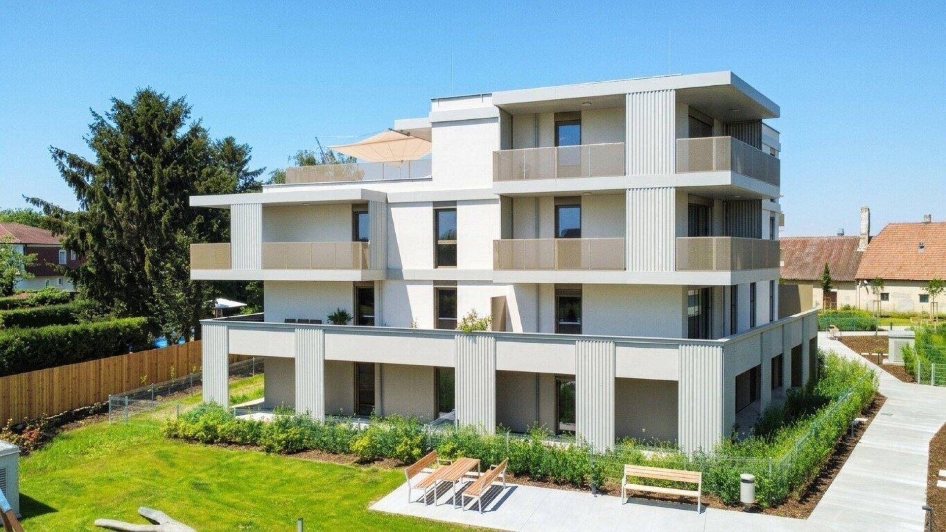 Modernes Wohngebäude mit Balkonen, einer Dachterrasse und einer kleinen Außensitzgruppe im Grünen.