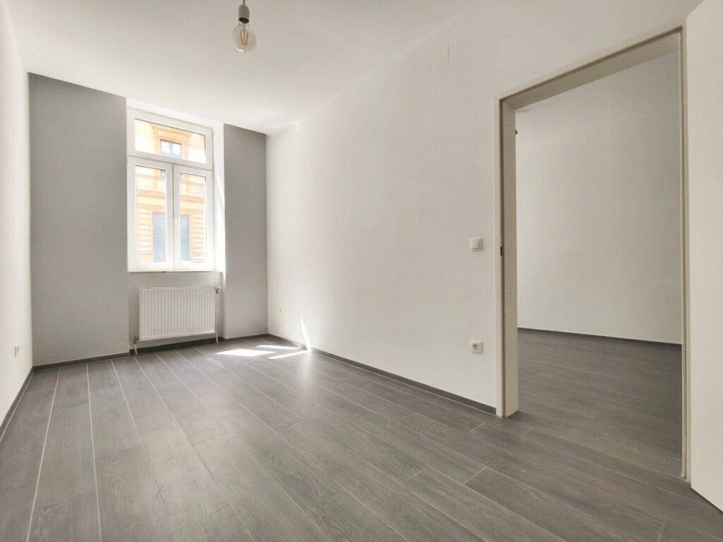 Lichtdurchfluteter Raum mit grauem Laminatboden, Fenster und einer grauen Akzentwand.