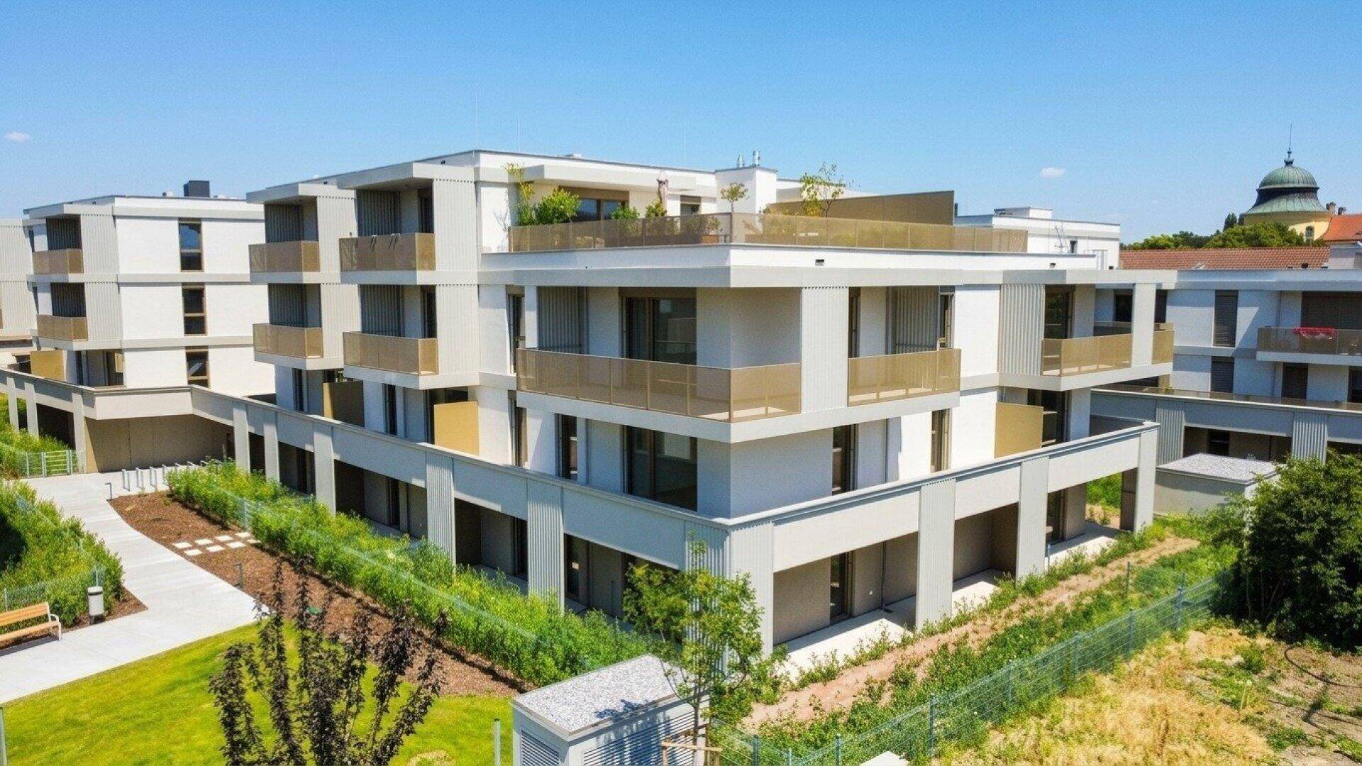 Modernes Wohngebäude mit Balkonen und einer Dachterrasse, umgeben von gepflegten Grünflächen unter blauem Himmel.