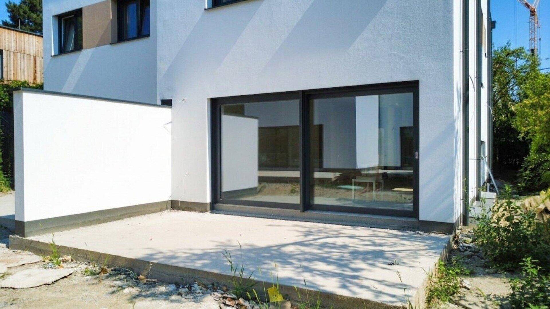Unmöblierte Terrasse am Neubau mit großen Schiebefenstern und direktem Zugang zum Gartenbereich.