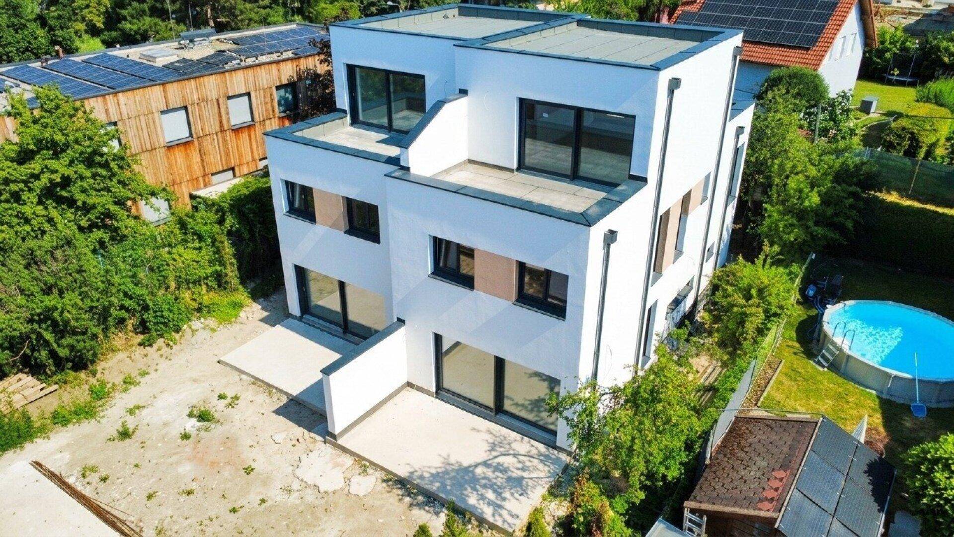 Luftaufnahme eines modernen Neubaus mit Flachdach und umliegenden Häusern in grüner Umgebung.