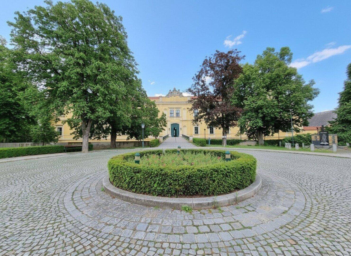 Historisches Gebäude mit gepflastertem Vorplatz, großen Bäumen und einem runden Blumenbeet.