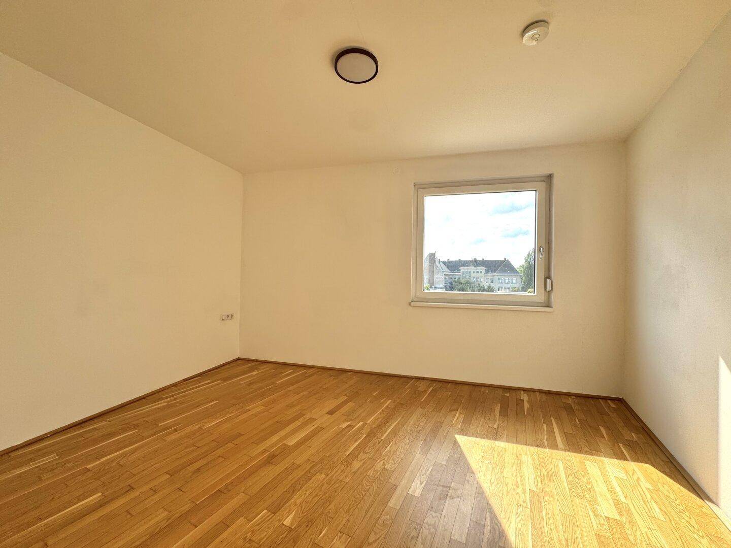 Helles Schlafzimmer mit Parkettboden und einem Fenster mit Blick auf die Umgebung.