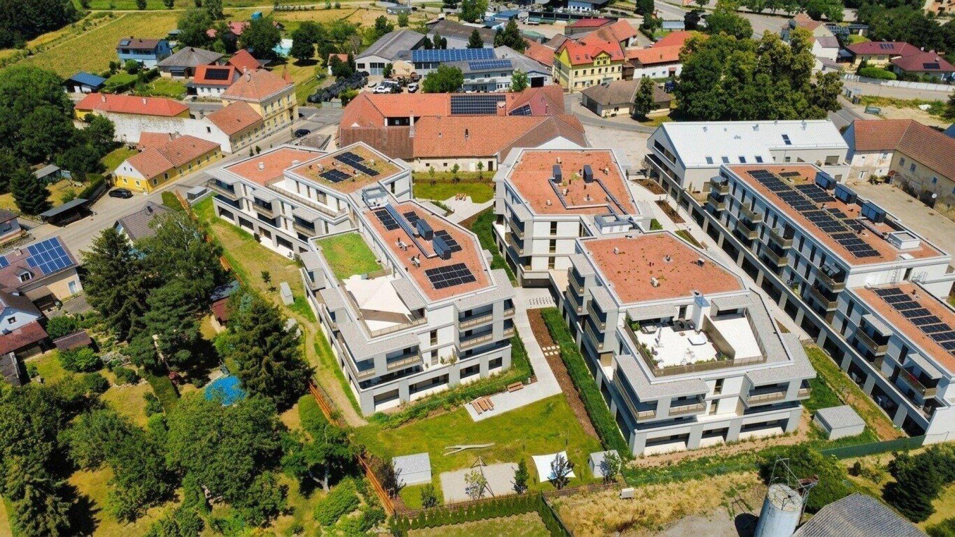 Luftaufnahme eines modernen Wohnkomplexes mit mehreren Gebäuden, Grünflächen und Photovoltaikanlagen.