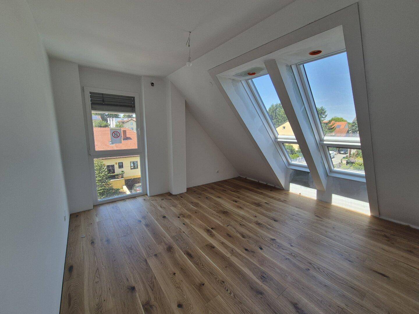 Lichtdurchfluteter Raum mit Holzboden und schrägen Dachfenstern, bietet einen Blick ins Grüne.