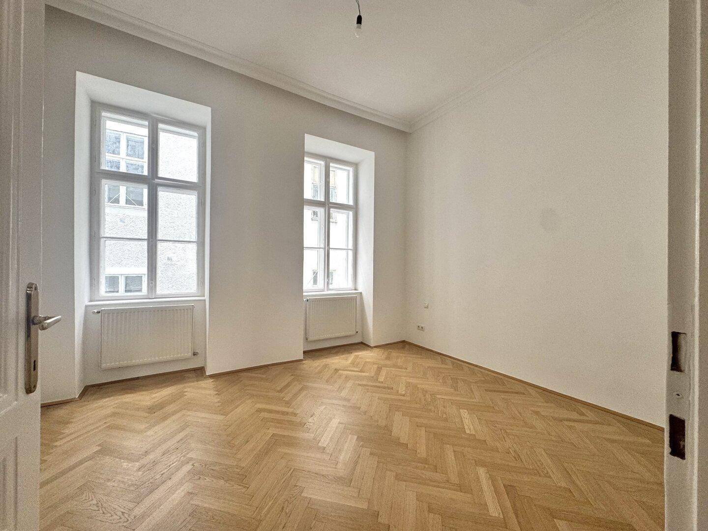 Helles Zimmer mit zwei Fenstern und klassischem Fischgrätparkett, bereit zur individuellen Gestaltung.