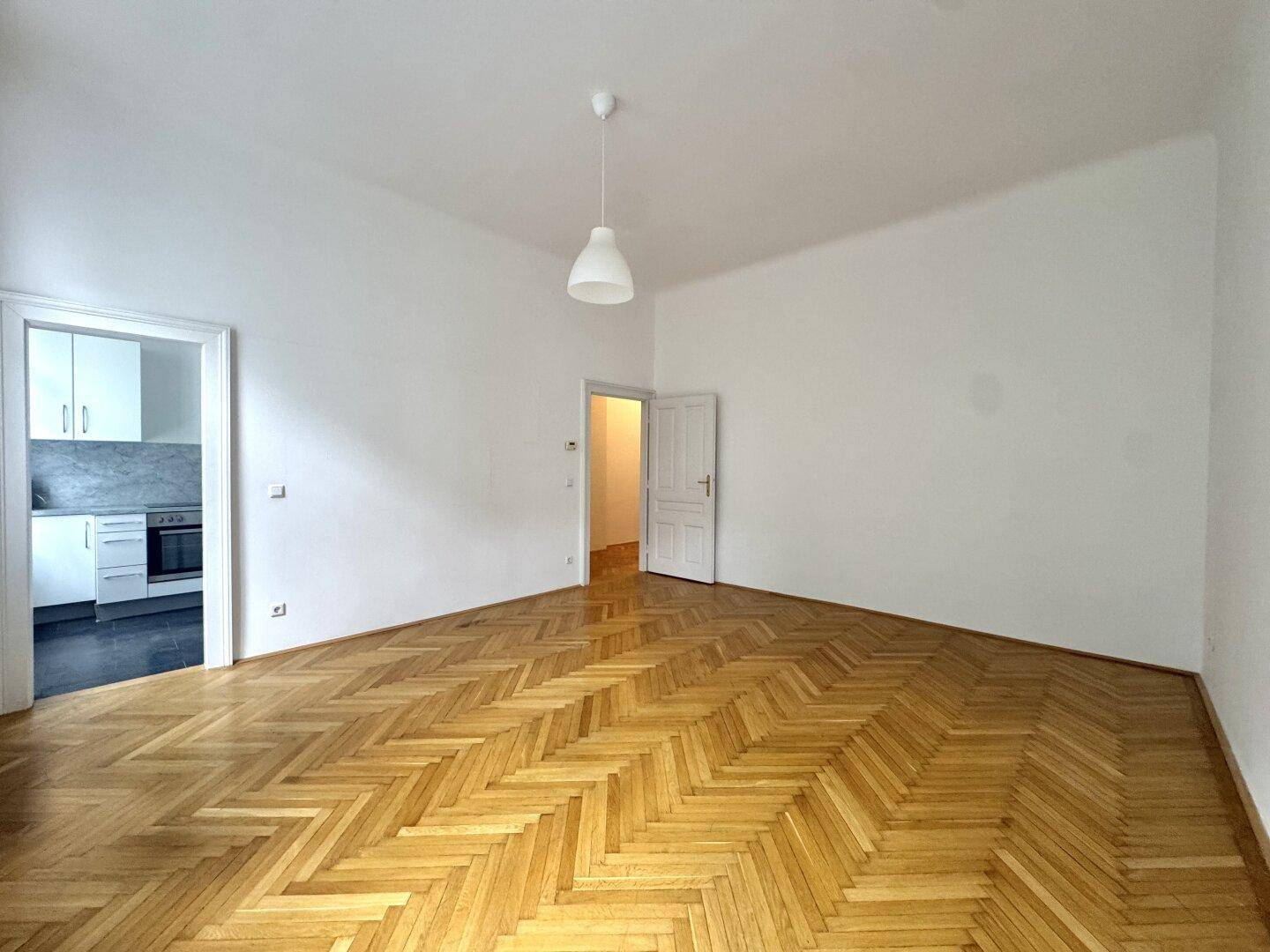 Heller Wohnbereich mit Fischgrätparkett und Zugang zu Küche und weiterem Zimmer.