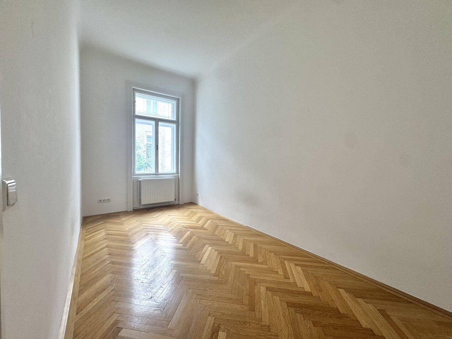 Ruhiges Zimmer mit Fischgrätparkett und einem großen Fenster, ideal als Schlaf- oder Arbeitszimmer.