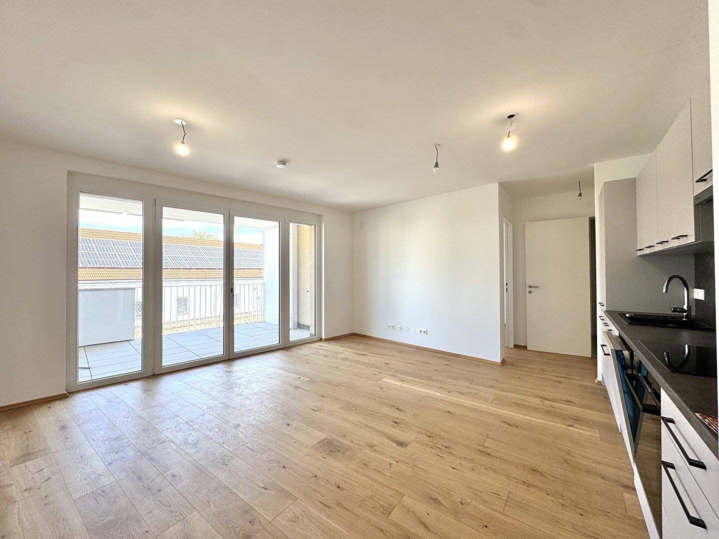Heller, leerer Wohnbereich mit Holzboden, großen Fenstern und Zugang zu einem Balkon.