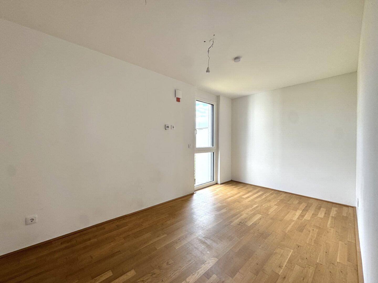 Leeres Zimmer mit Holzboden und einem hohen Fenster, ideal als Schlaf- oder Arbeitszimmer.