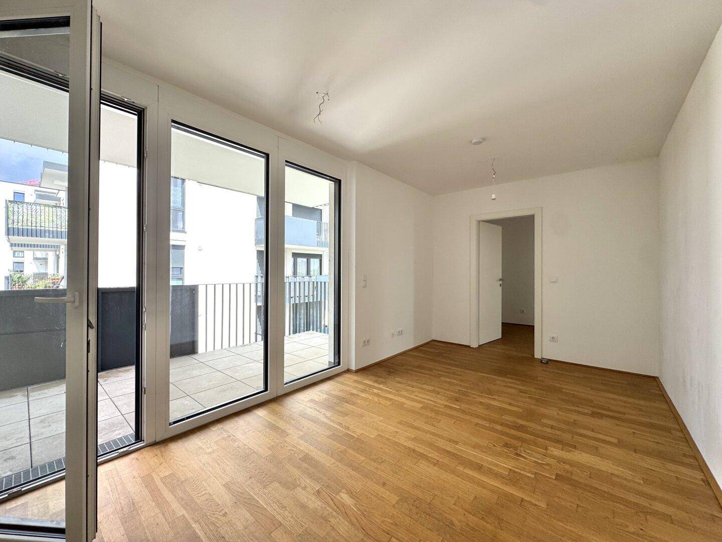 Heller Wohnbereich mit Holzboden und großen Fenstern, die Zugang zu einem Balkon bieten.