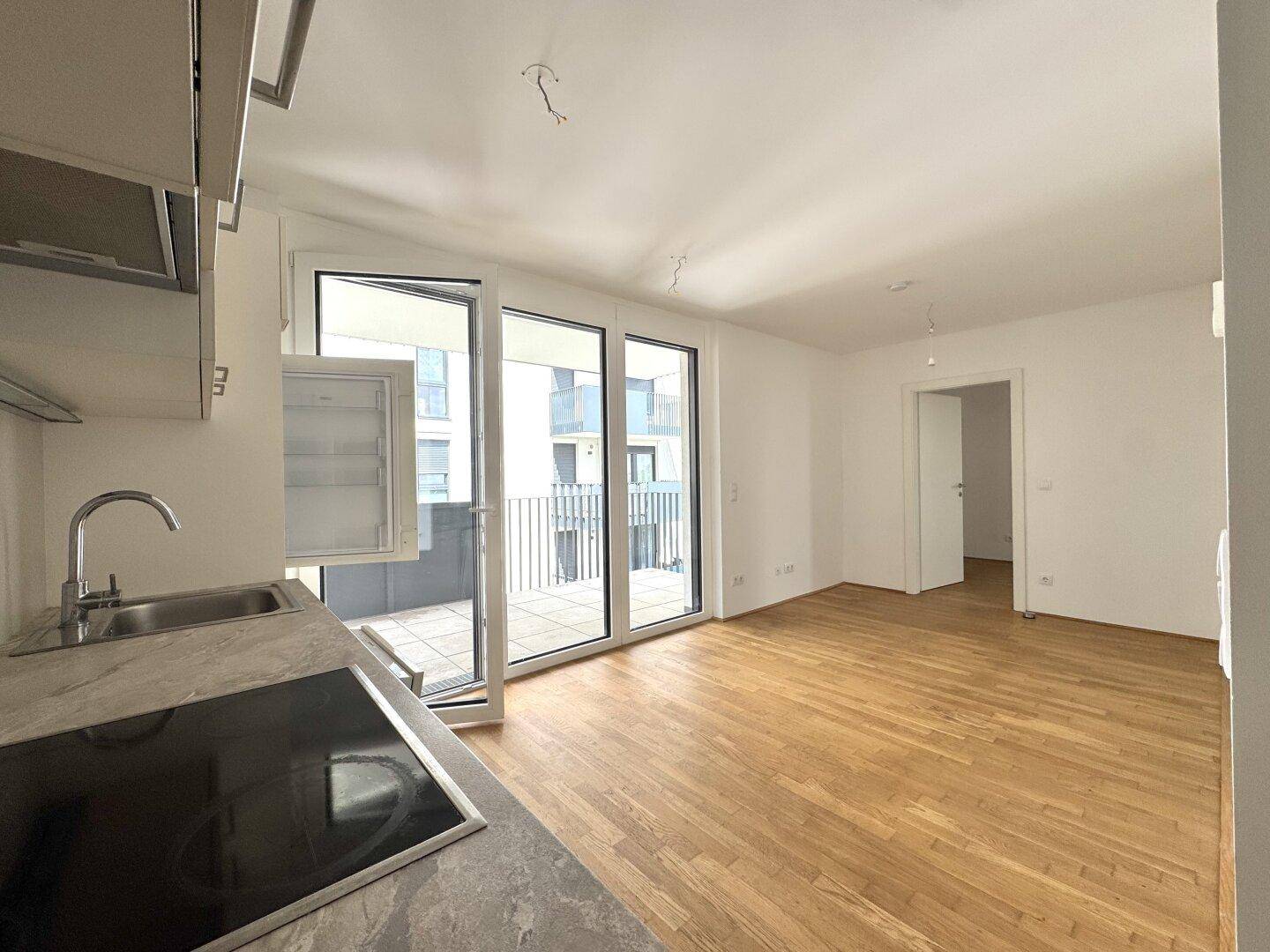 Offener Wohnbereich mit Holzboden und Küchenzeile, direktem Zugang zum Balkon durch große Glasfenster.