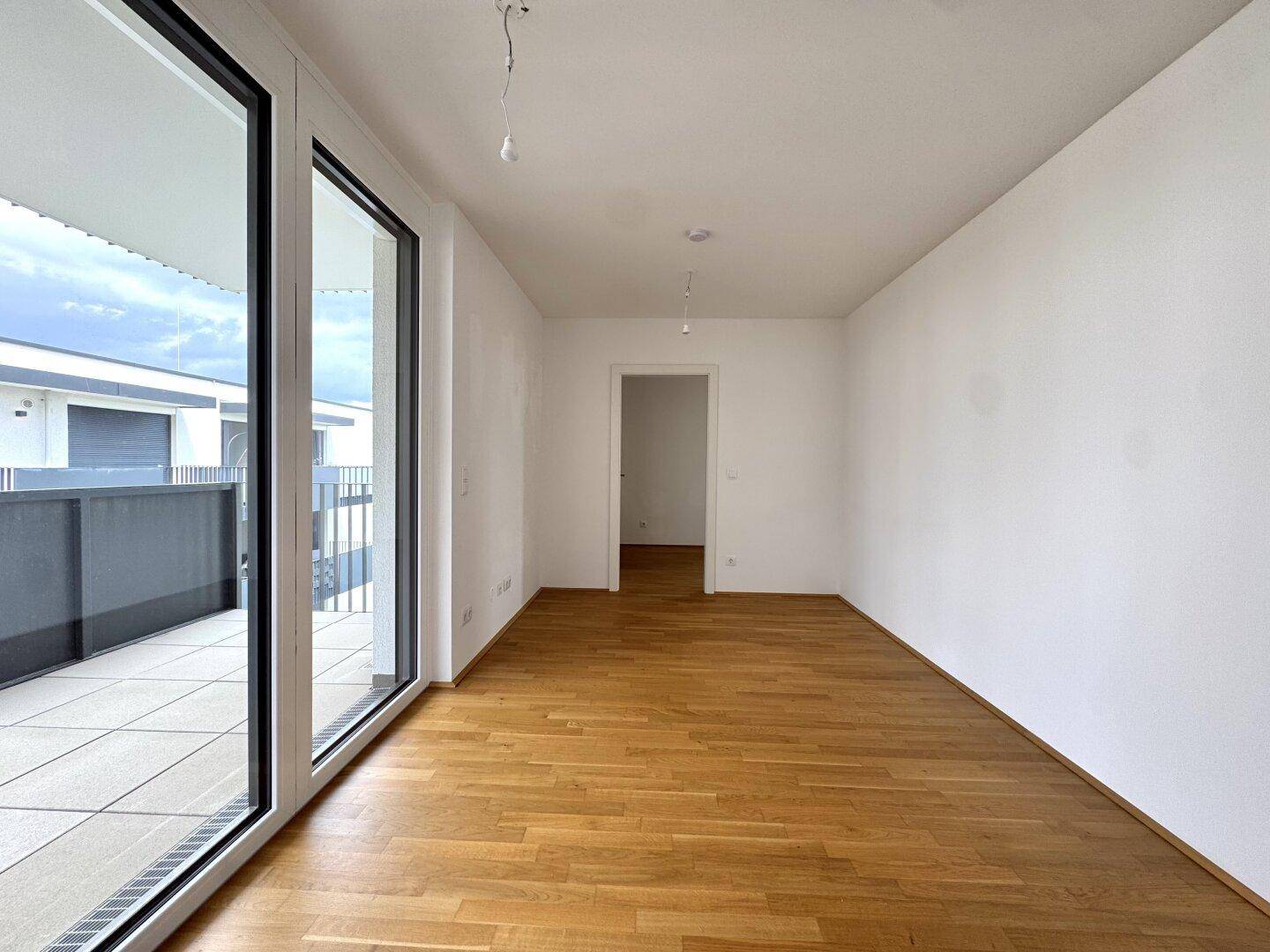 Heller, leerer Raum mit Holzparkettboden und großen Fenstern zum Balkon.