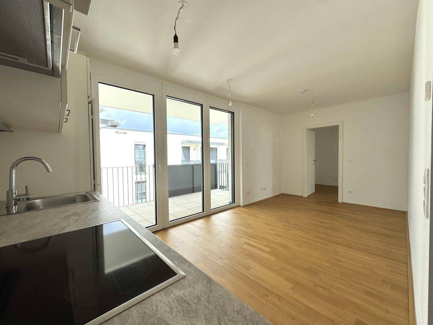 Offener Wohnbereich mit Kochnische, Holzparkett und Zugang zu einem Balkon.