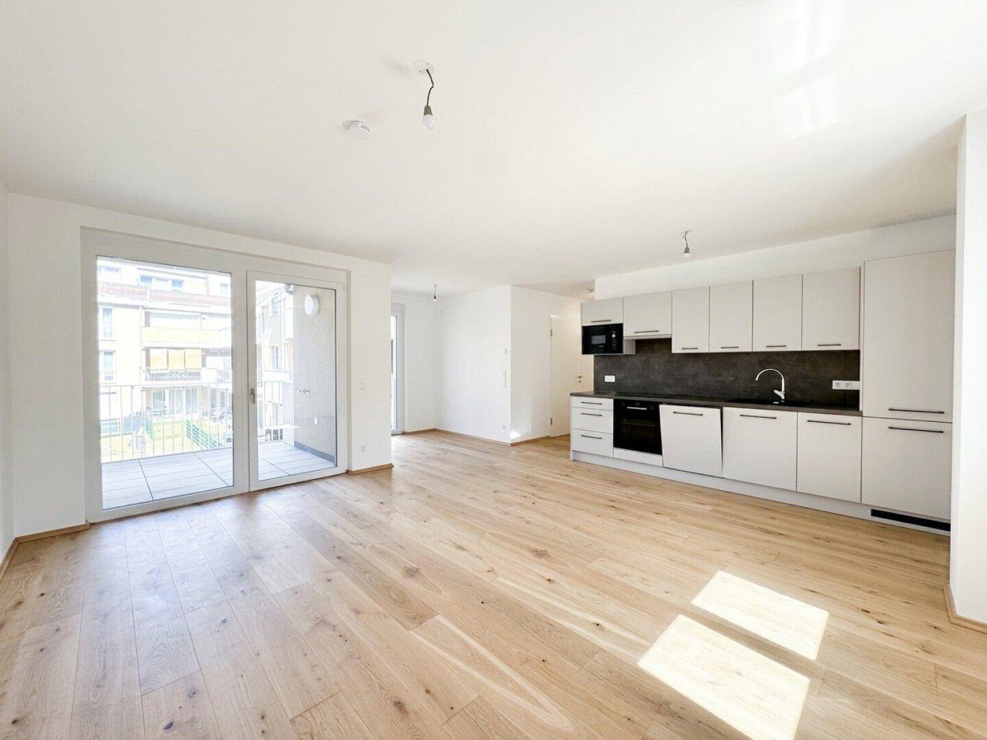 Heller offener Wohnbereich mit moderner Einbauküche, Holzboden und Zugang zum Balkon.