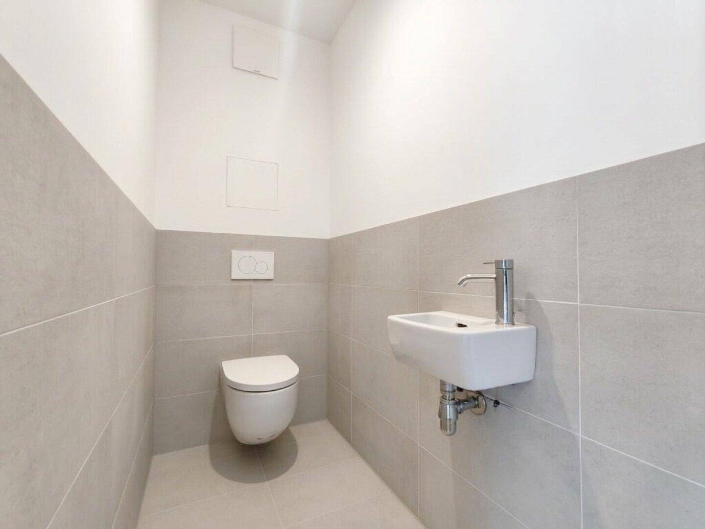 Separates Gäste-WC mit Wand-WC, kleinem Waschbecken und modernen grauen Fliesen.