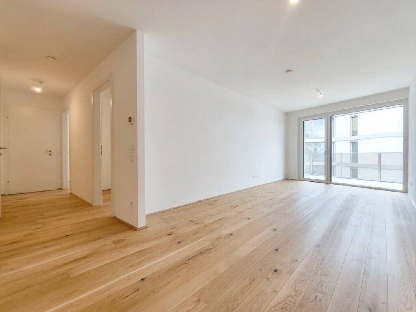 Geräumiges, leeres Wohnzimmer mit hellem Holzboden und Zugang zu einem Balkon.