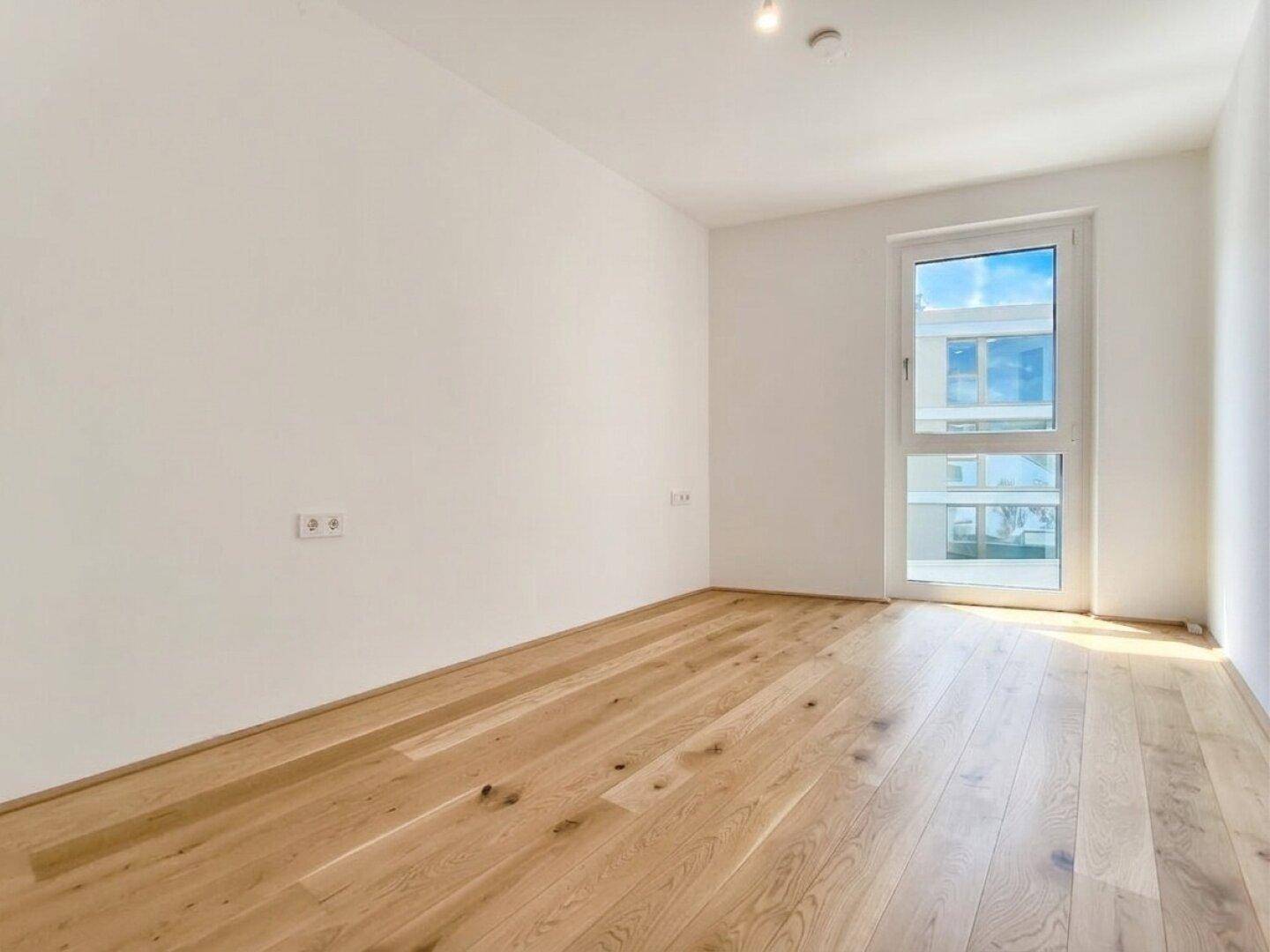 Leeres Schlafzimmer mit Holzboden und einem großen Fenster, das den Raum erhellt.