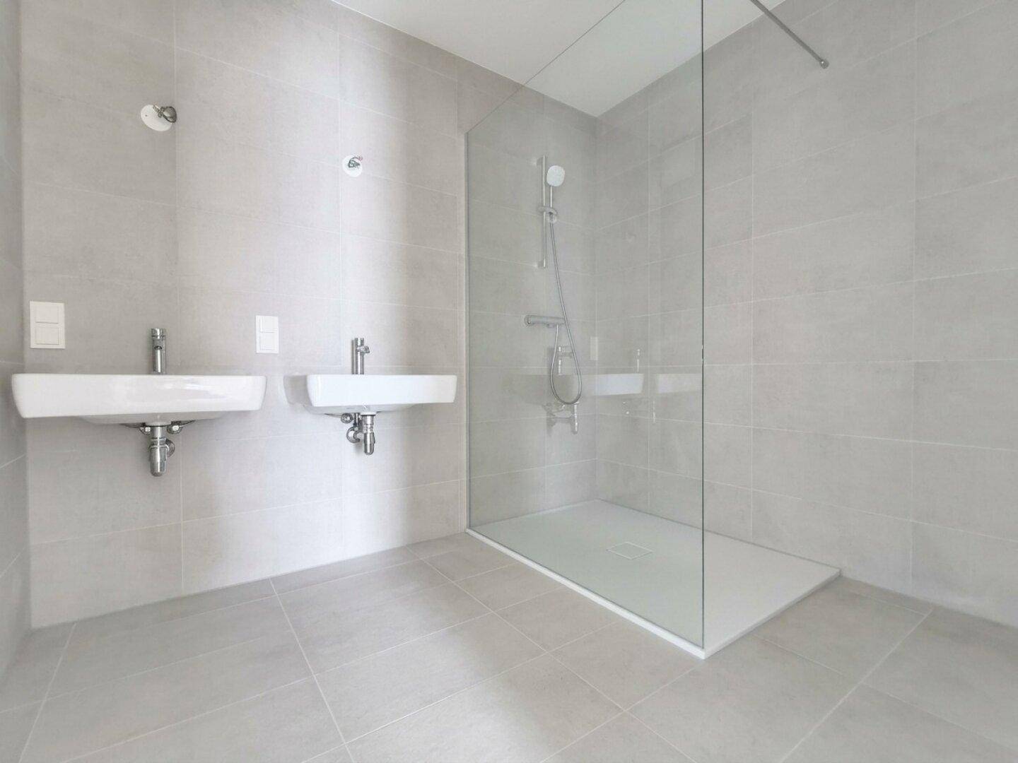 Modernes Badezimmer mit hellgrauen Fliesen, zwei Waschbecken und einer begehbaren Dusche.