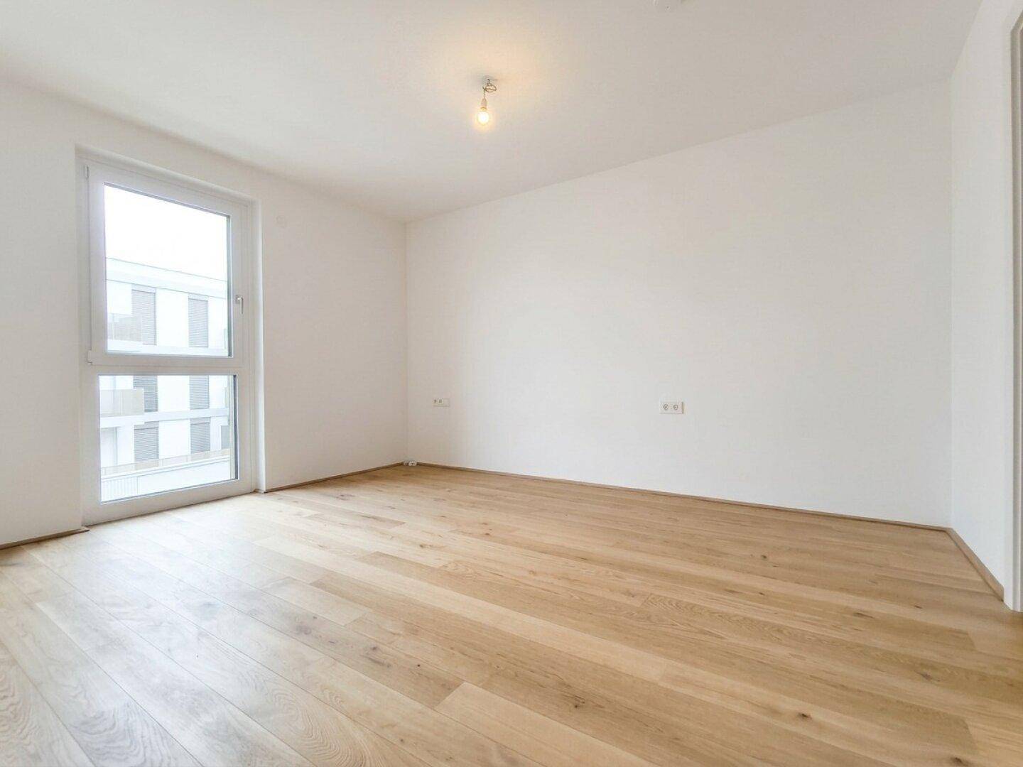 Geräumiges Zimmer mit Parkettboden und Fenster, das viel Tageslicht hereinlässt.