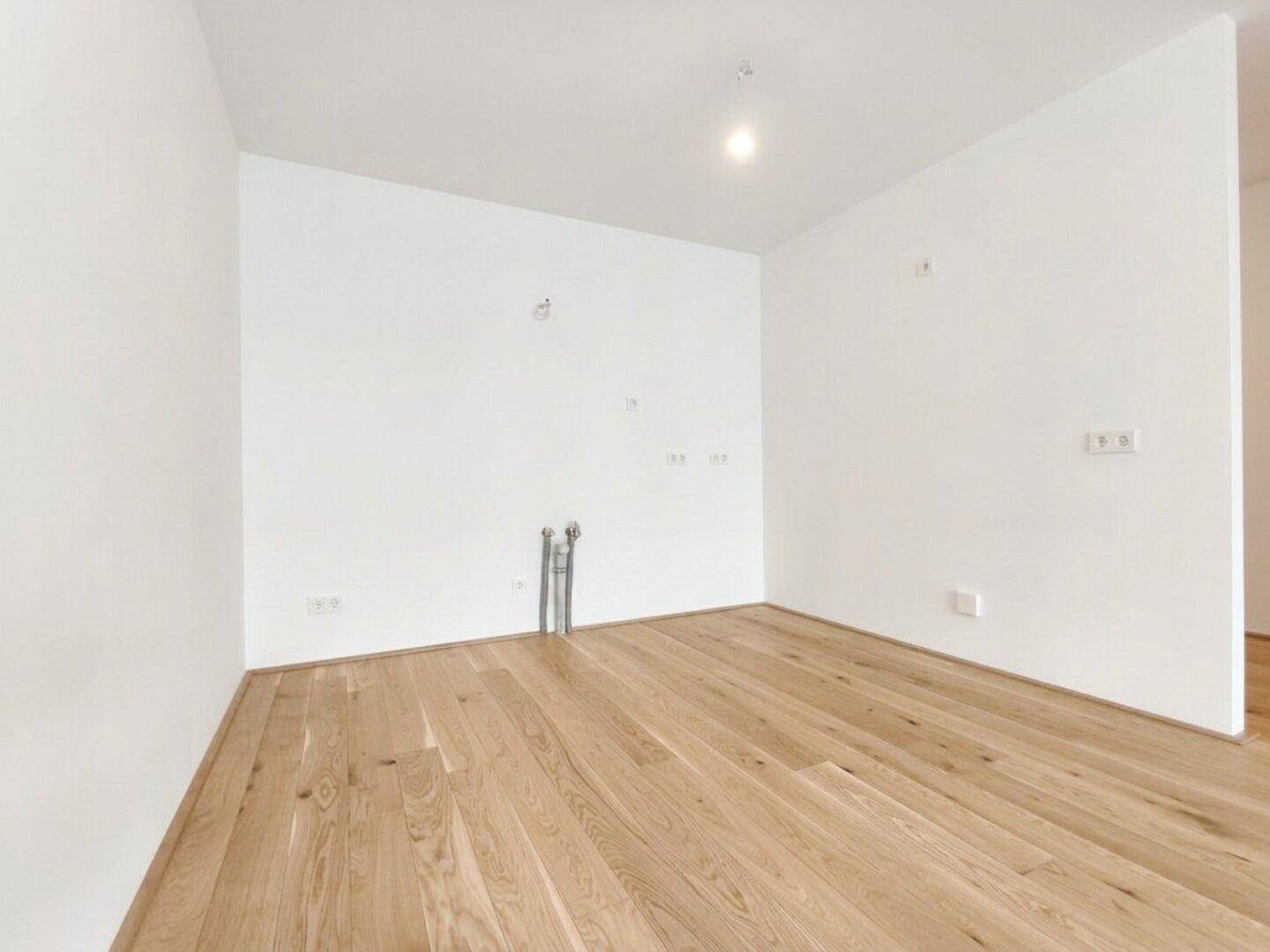 Leerer Küchenbereich mit Anschlüssen und hellem Holzparkett, bereit für den Einbau einer modernen Küche.