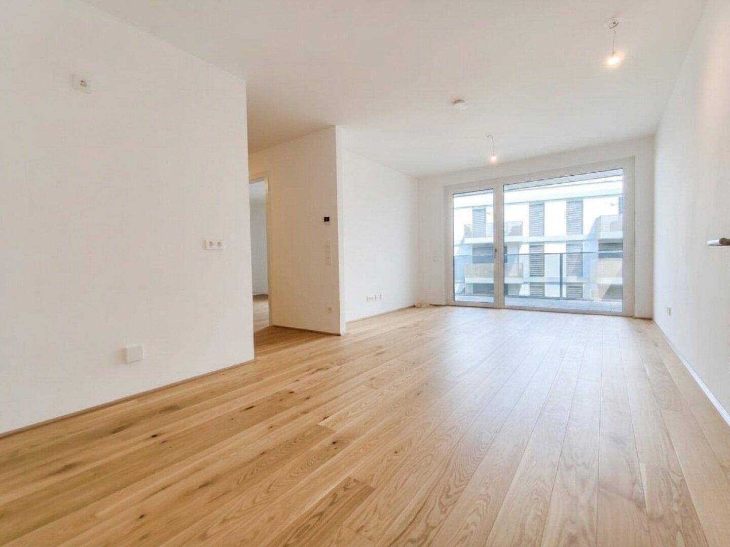Helles, leeres Zimmer mit hellem Holzparkett und Zugang zu einem Balkon, ideal als Wohnbereich.