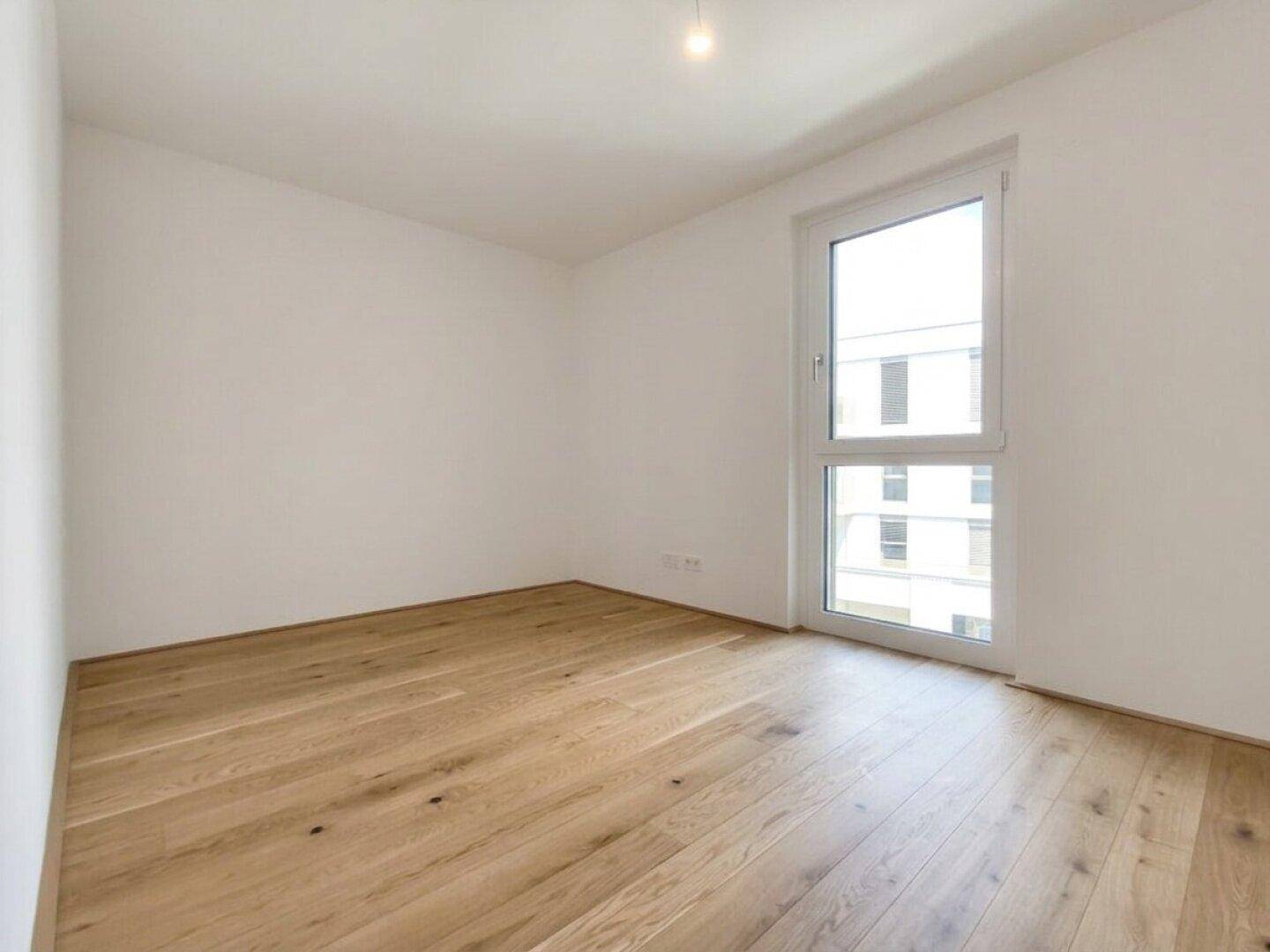 Helles Schlafzimmer mit Holzparkett und einem großen Fenster, das viel Tageslicht hereinlässt.