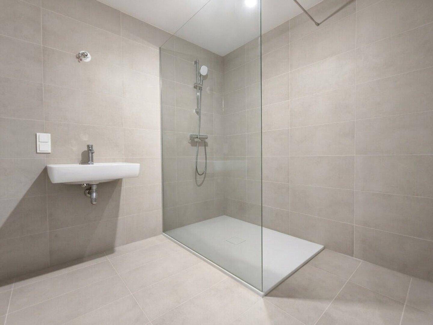 Modernes Badezimmer mit grauen Fliesen, wandhängendem Waschbecken und einer geräumigen begehbaren Dusche.