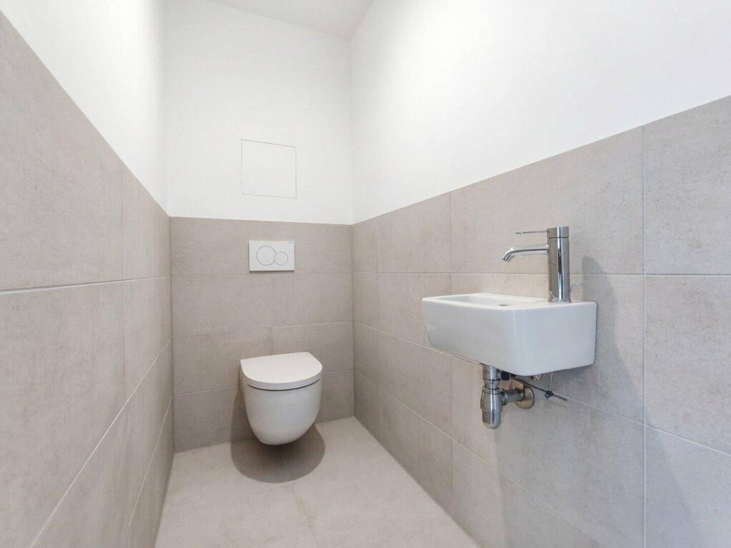 Modernes Gäste-WC mit grauen Fliesen, wandhängendem WC und kleinem Waschbecken.