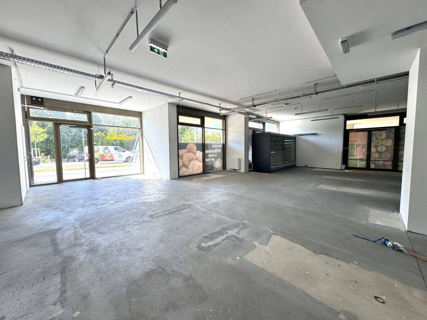 Geräumiger, leerer Ladenbereich mit großen Schaufenstern, Betonboden und freiliegenden Deckeninstallationen.